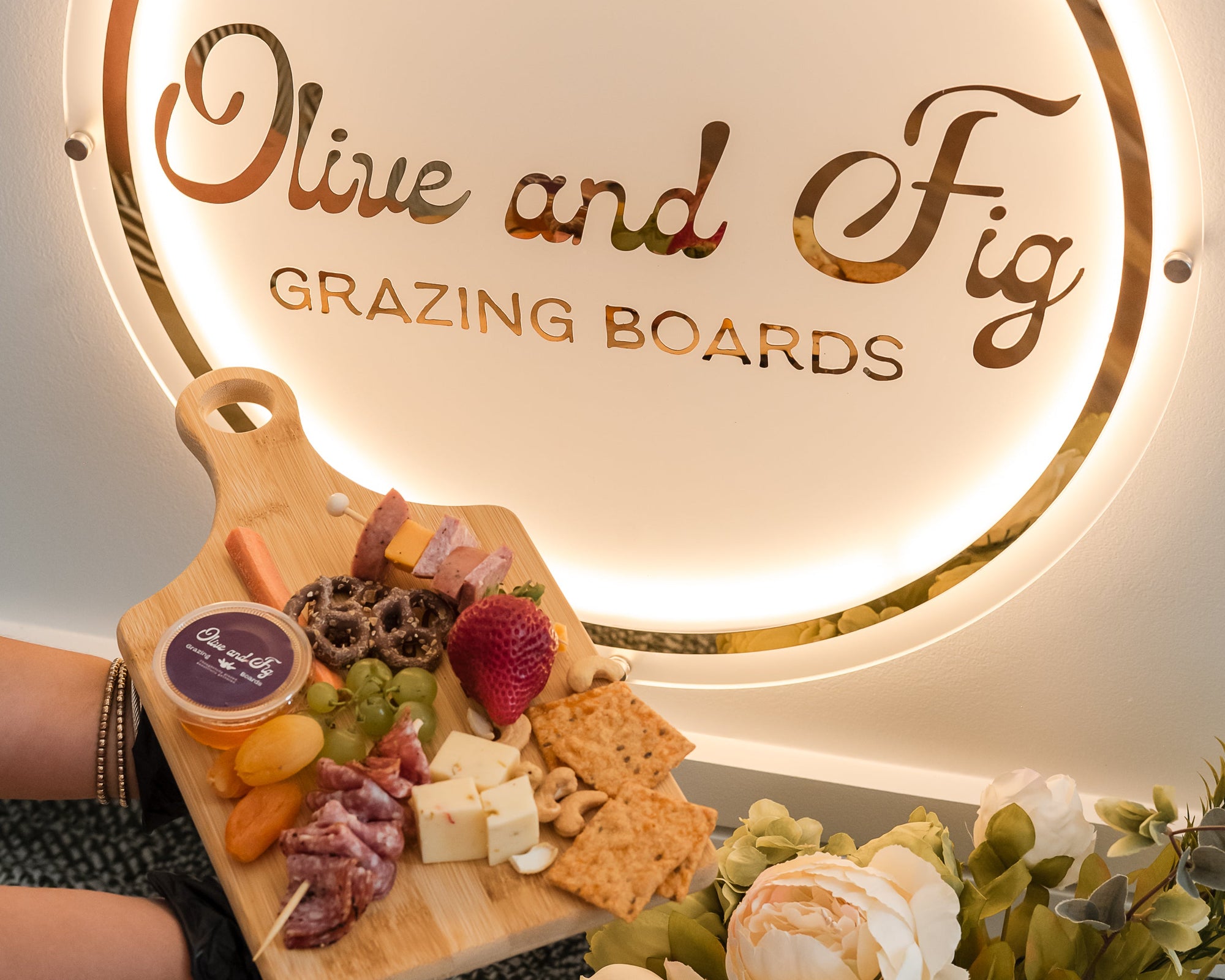 Grazing board with assorted foods in front of 'Olive and Fig Grazing Boards' sign.